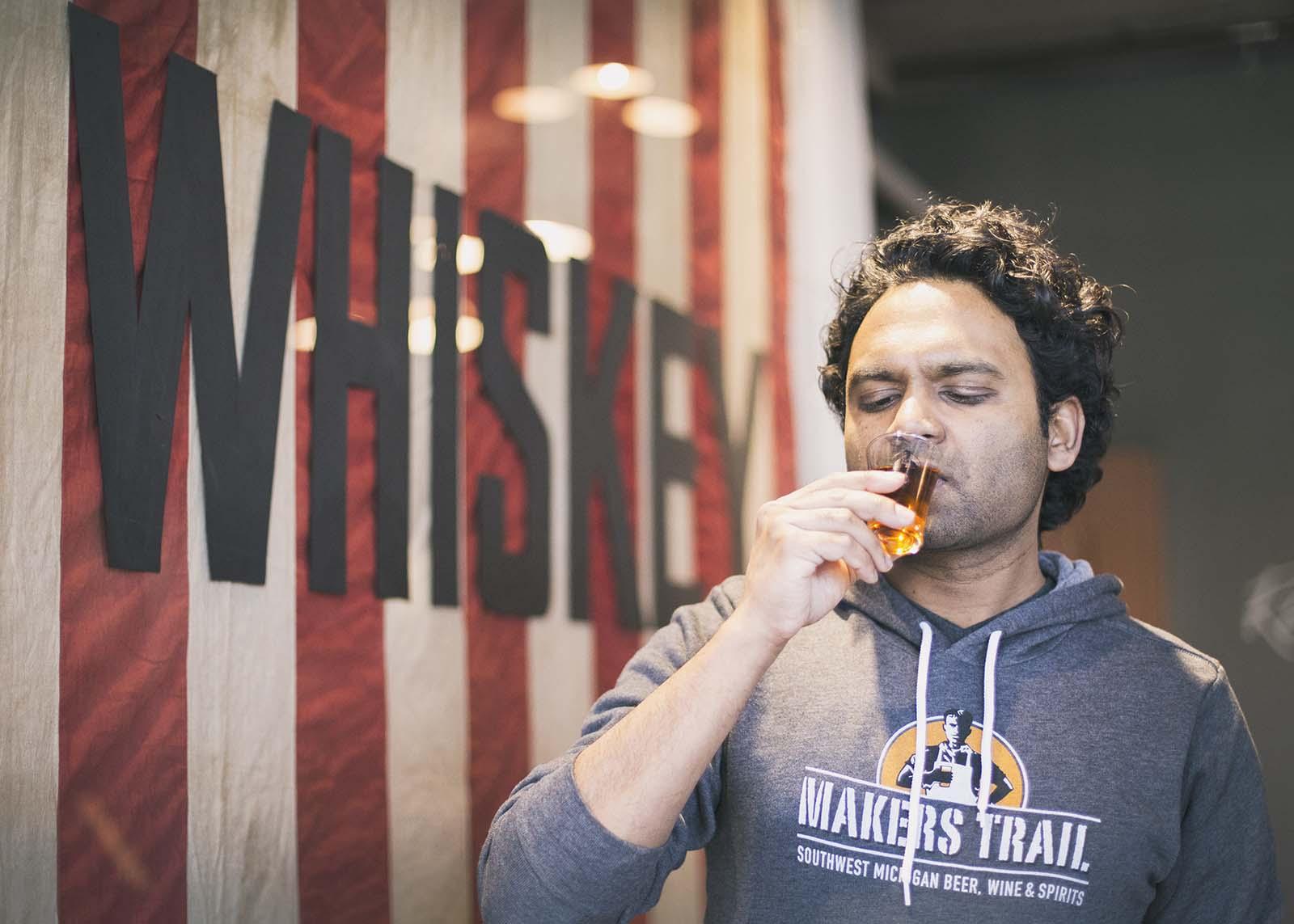 Man drinking whiskey in front of the whiskey flag at Journeyman Distillery