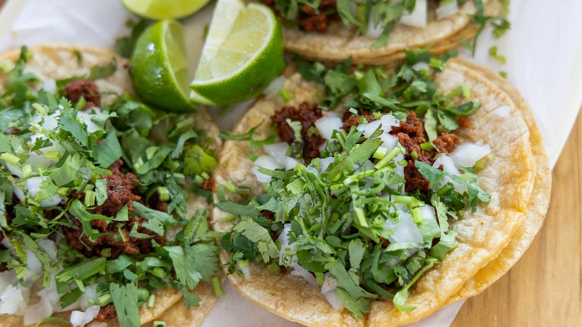 A close-up of three al pastor tacos with limes nestled between them, served on a plate 