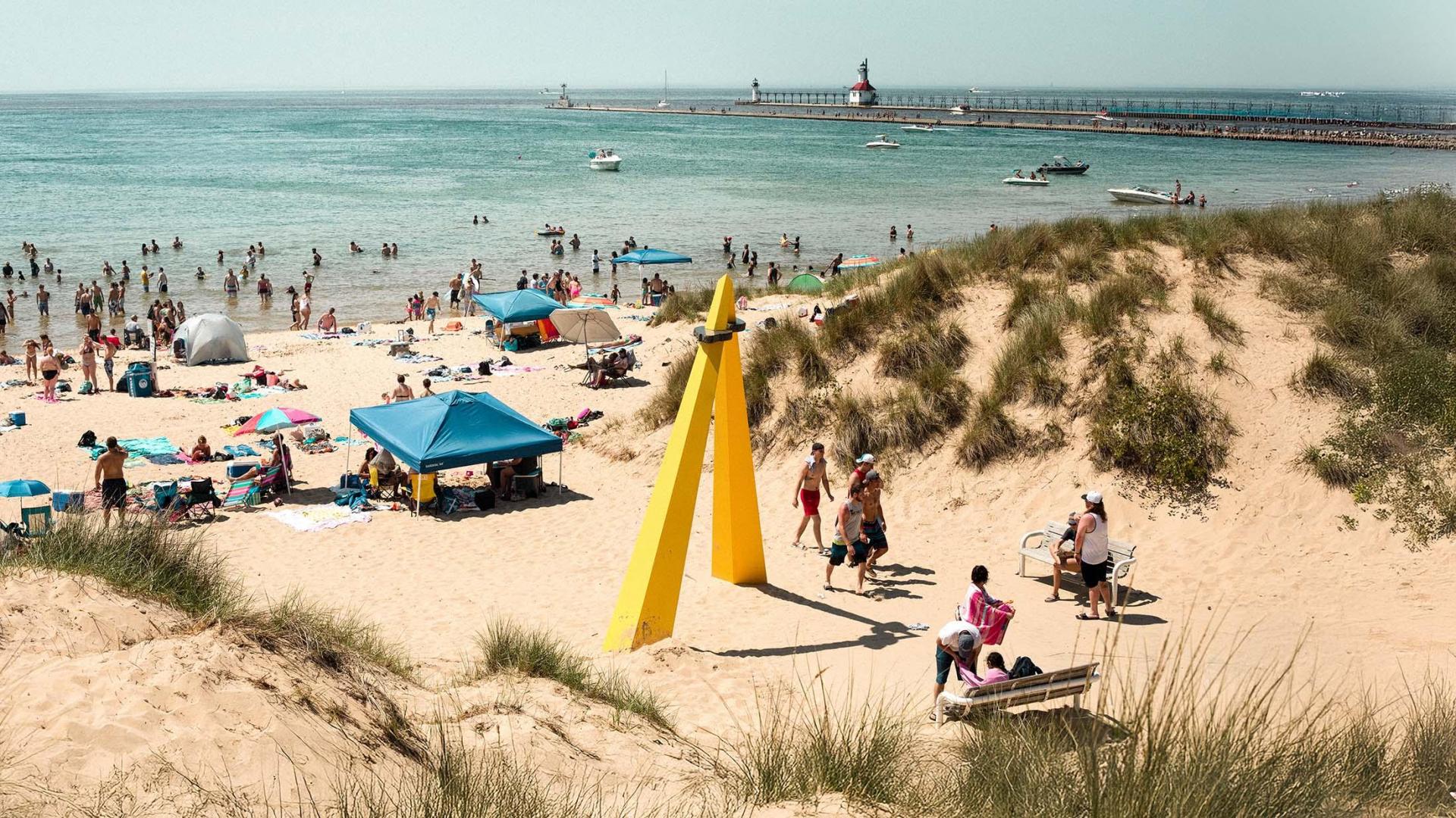 A wide shot of Silver Beach on a busy, sunny summer day, with people sunbathing, playing sports, and swimming in Lake Michigan, and the North Pier Lighthouse visible in the background