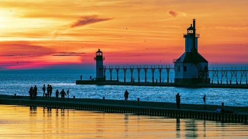 People walking on the South Pier during a Lake Michigan sunset, with the North Pier Lighthouse in the background and the sky glowing in warm orange and pink hues