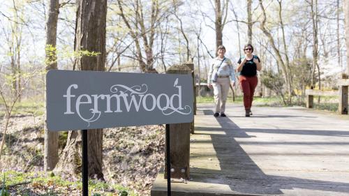 Two women walking over a wooden bridge toward a sign for Fernwood Botanical Garden, surrounded by lush greenery