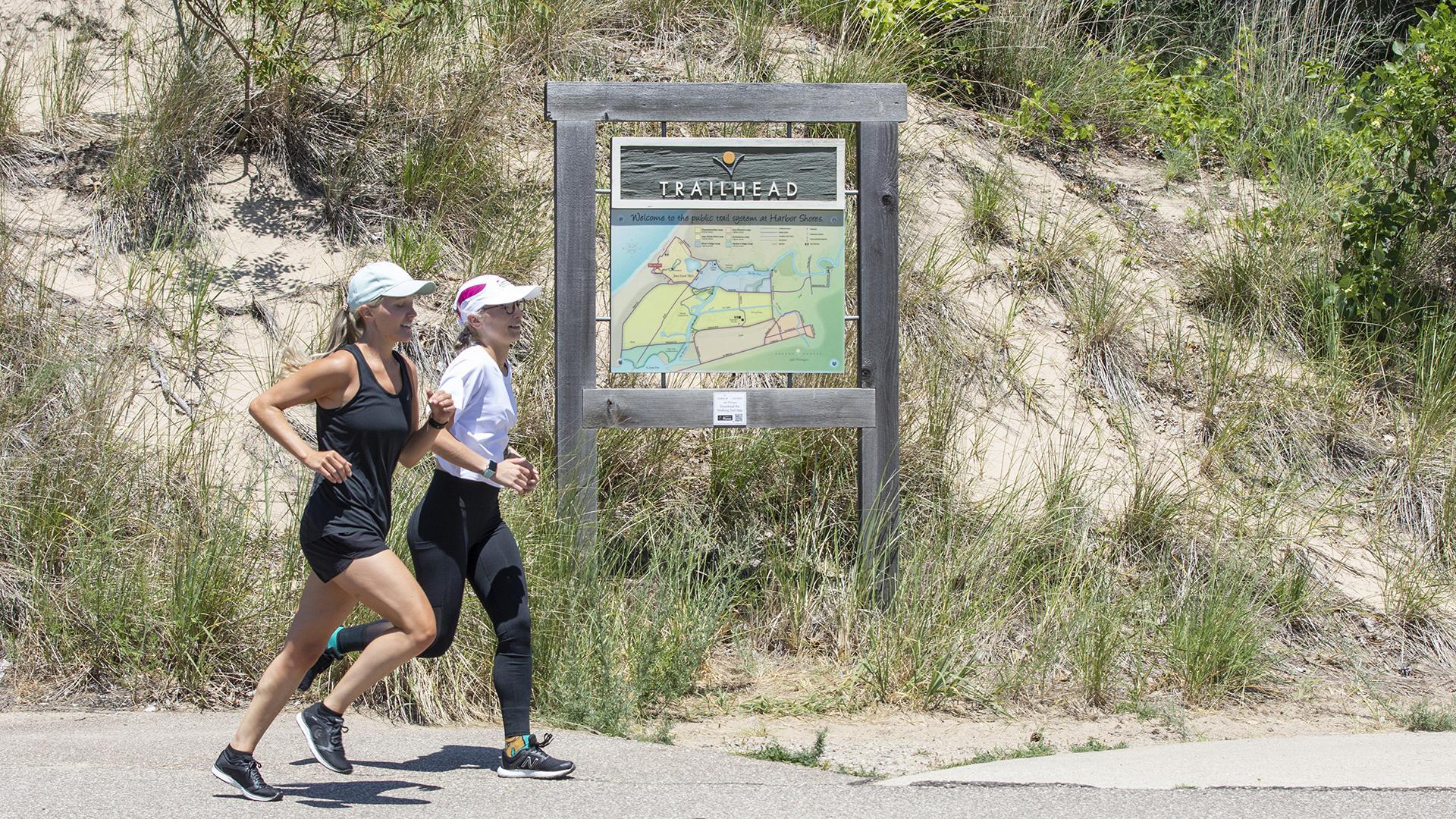 Two women running on a trail at Harbor Shores, with sand and dune grass in the background