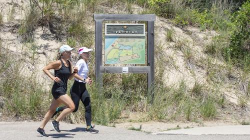 Two women running on a trail at Harbor Shores, with sand and dune grass in the background