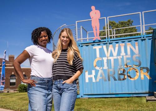 Two women smiling in front of an art sculpture and a blue shipping container transformed into a business with -Twin Cities Harbor- prominently displayed on the side