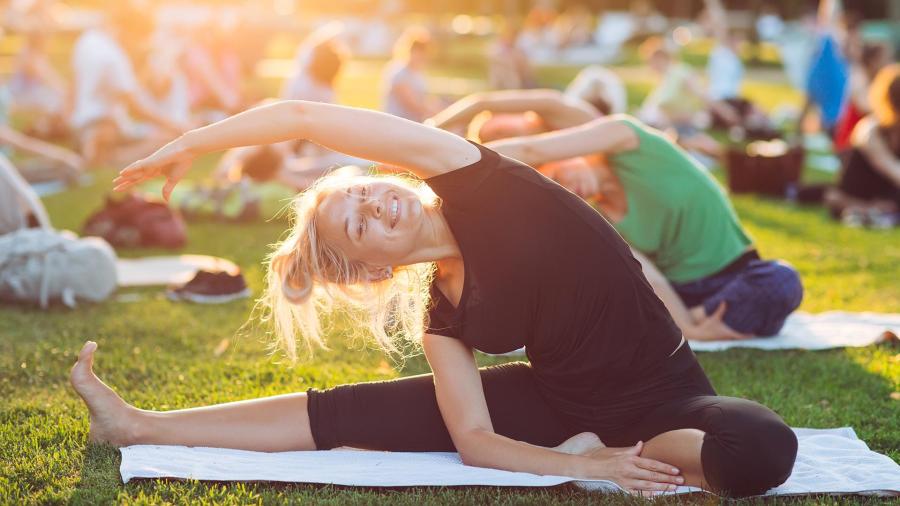 A woman smiling while practicing yoga outdoors, with a group of people practicing yoga in the background. The setting is peaceful and serene