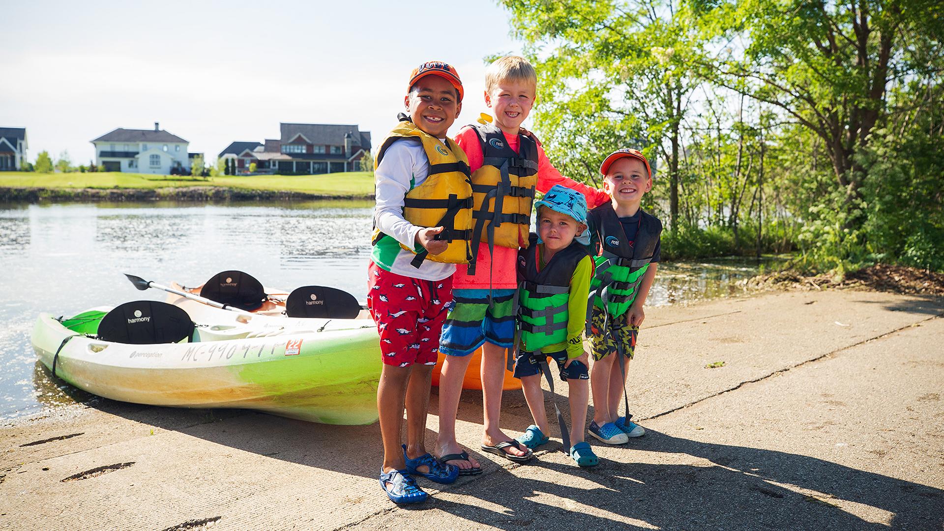 A group of kids in lifejackets smiling at the camera with kayaks behind them, excited and ready to go kayaking