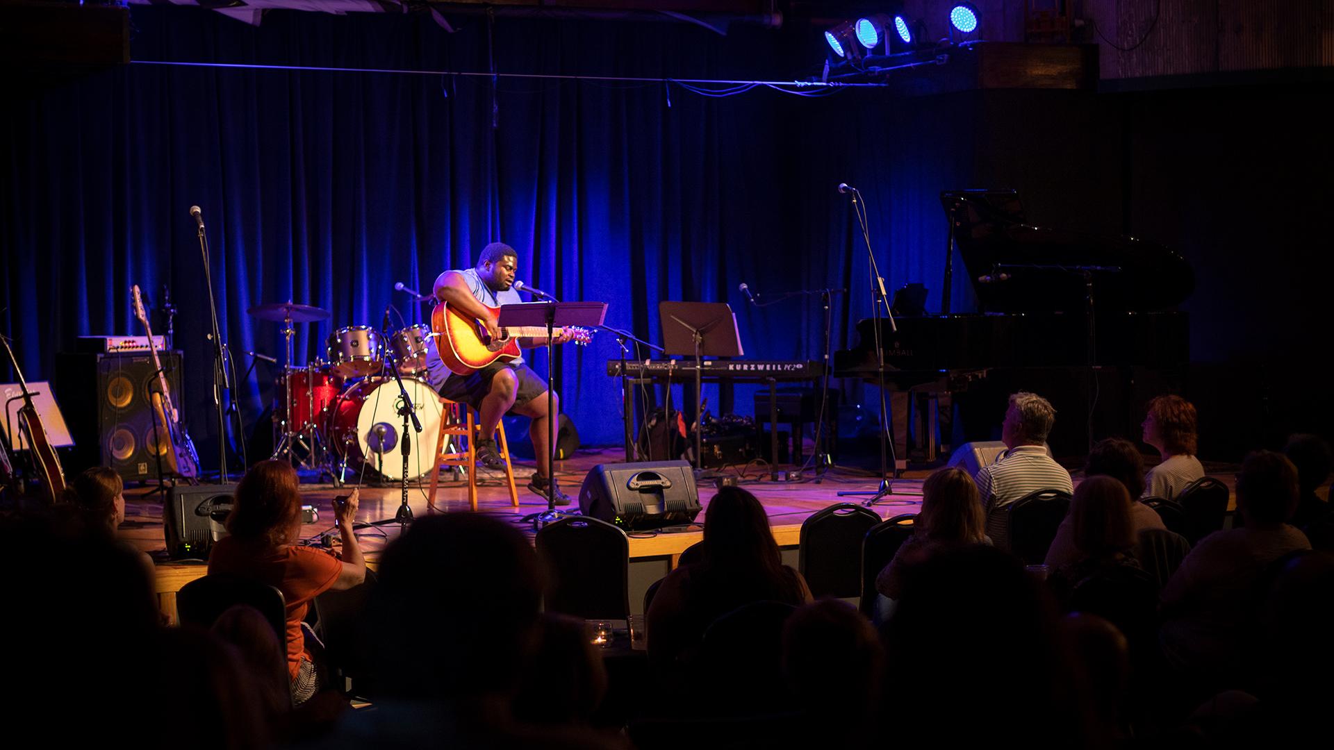 A man performing with his guitar for an audience at the Acorn Theater