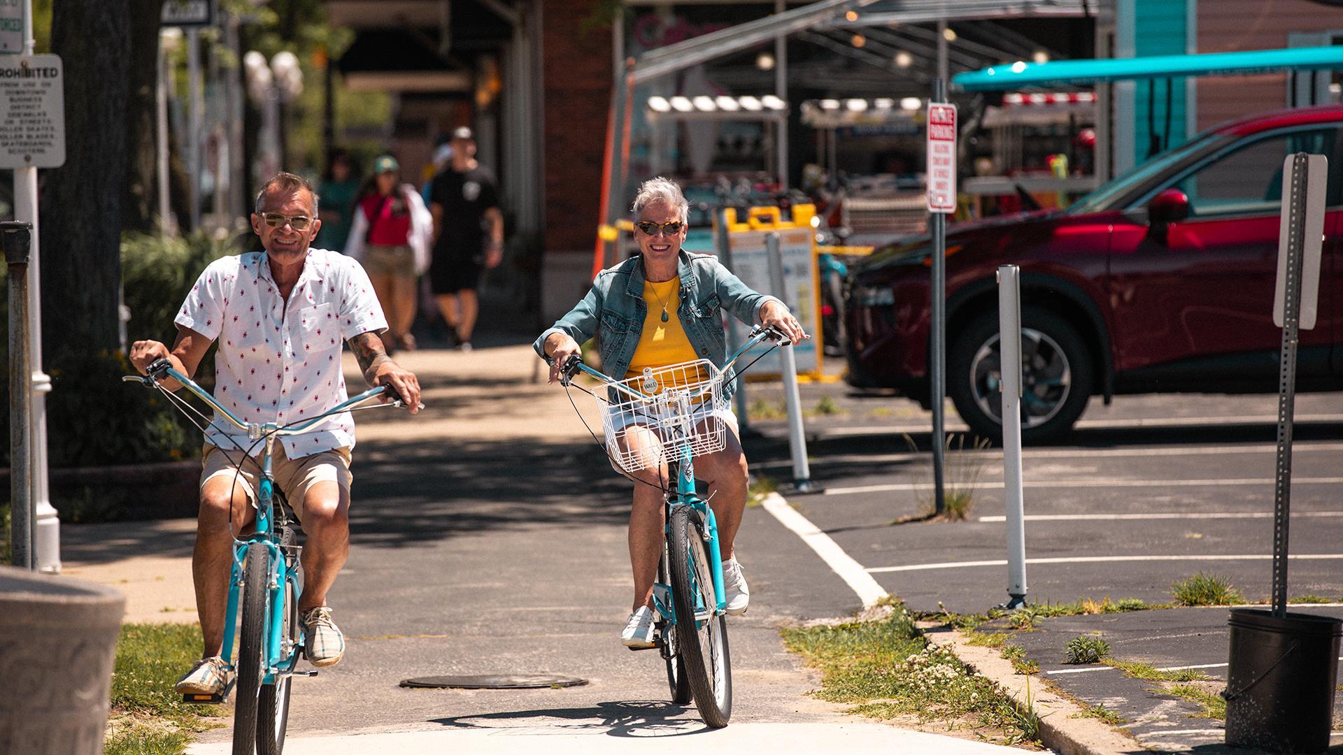An older couple riding surrey bikes through downtown St. Joe on a sunny day, smiling as they pedal past shops and trees along the charming streets