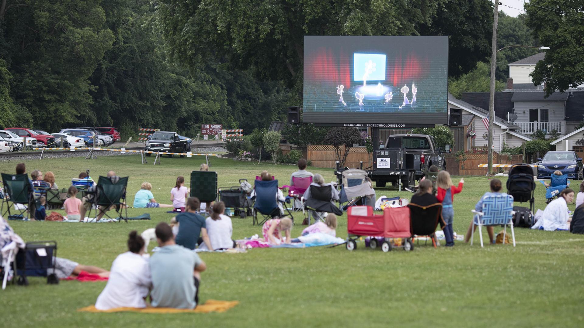 A summer outdoor movie on a grassy lawn, with people picnicking on blankets as they watch the screen