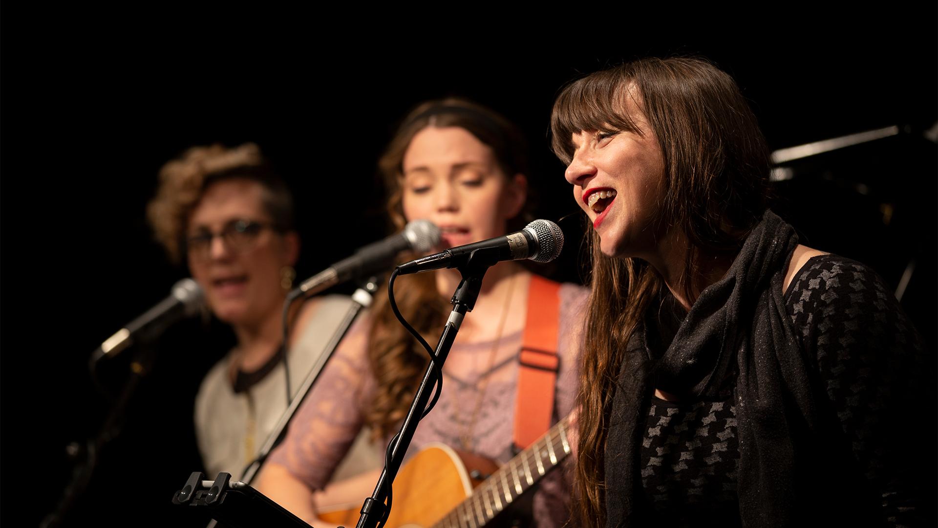 Three women singing at the Box Factory for the Arts, with the focus on the woman to the right as the main singer