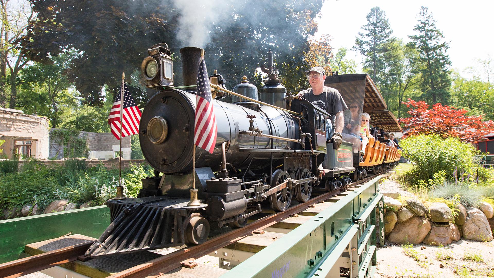 Miniature train at Eden Springs Park on a sunny day, with American flags on the front and children riding in the background