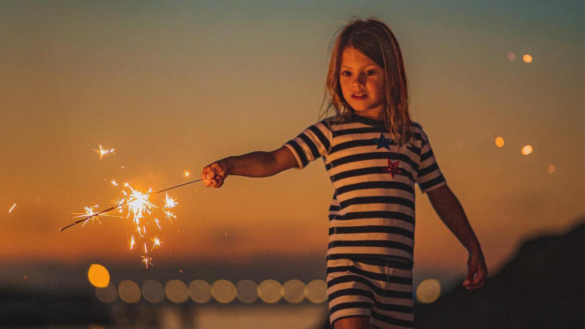 A young girl playing with sparklers on the beach as the sunset sky glows orange and the light begins to dim
