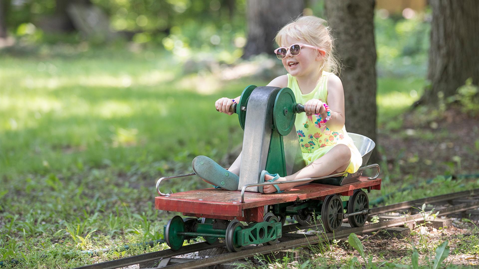 A young girl wearing sunglasses and a bright floral dress smiles while riding a small handcar on a miniature railroad track at Eden Springs Park