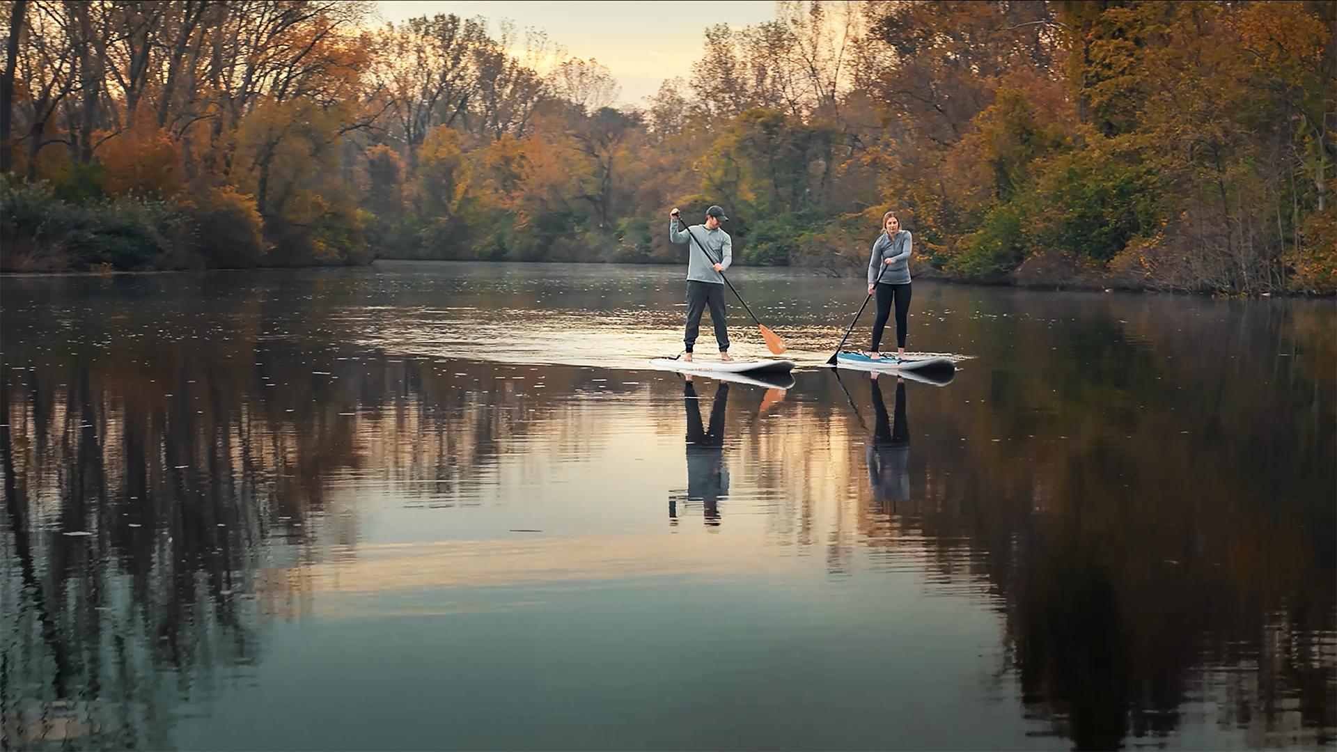 Two people paddleboarding side by side down a river on a fall day, surrounded by colorful autumn foliage
