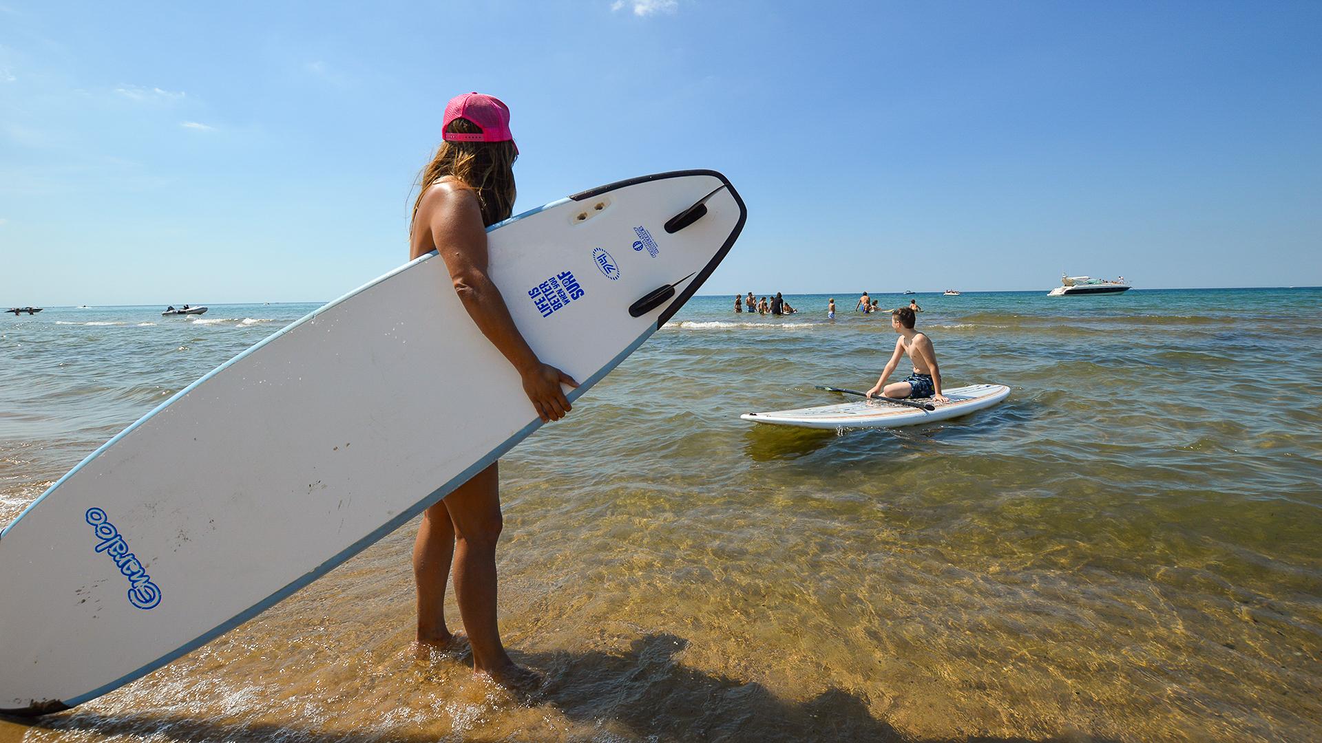 A close-up of a woman holding a paddleboard, preparing to join others on the water. In the background, a boy is already on a paddleboard and a group of people, some with boards, are gathered nearby