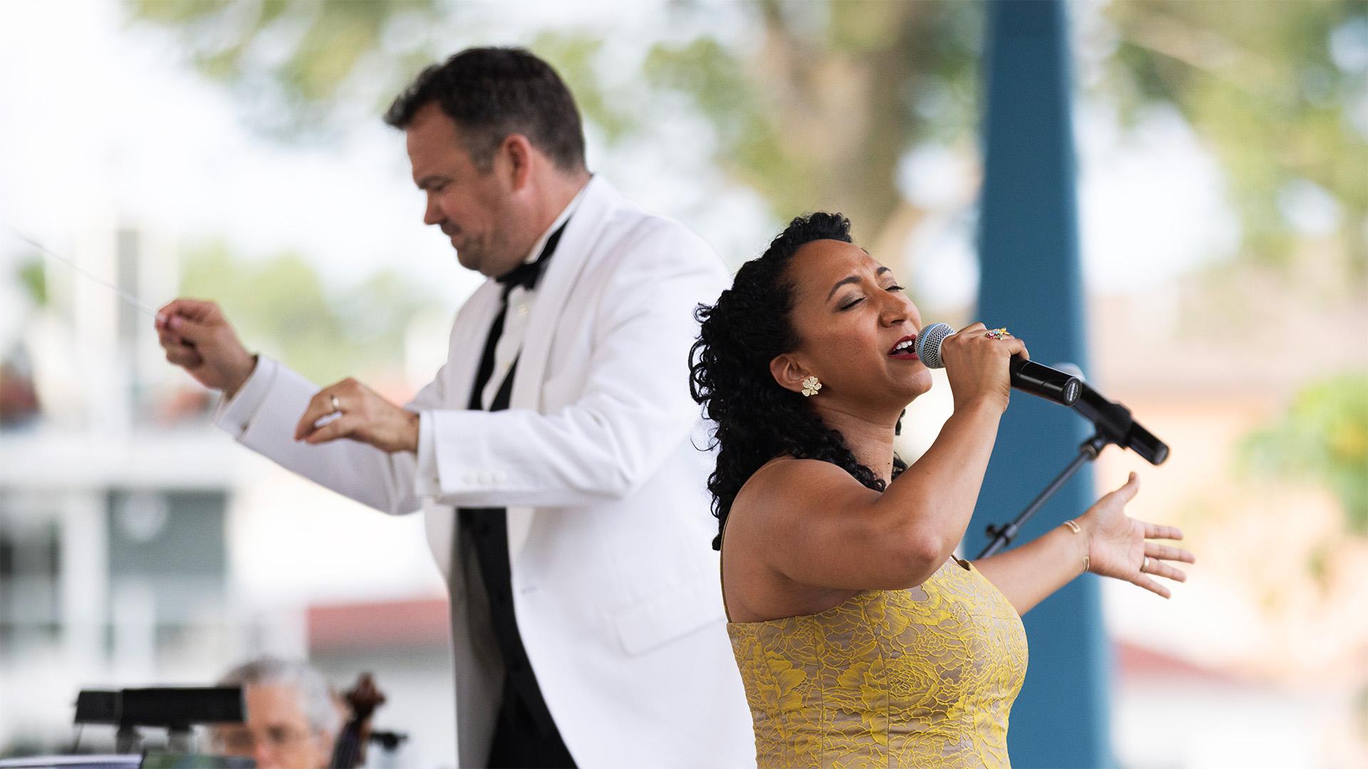 Outdoors at Shadowland Pavilion, a woman in a gold dress passionately sings on stage as a conductor leads the orchestra behind her.