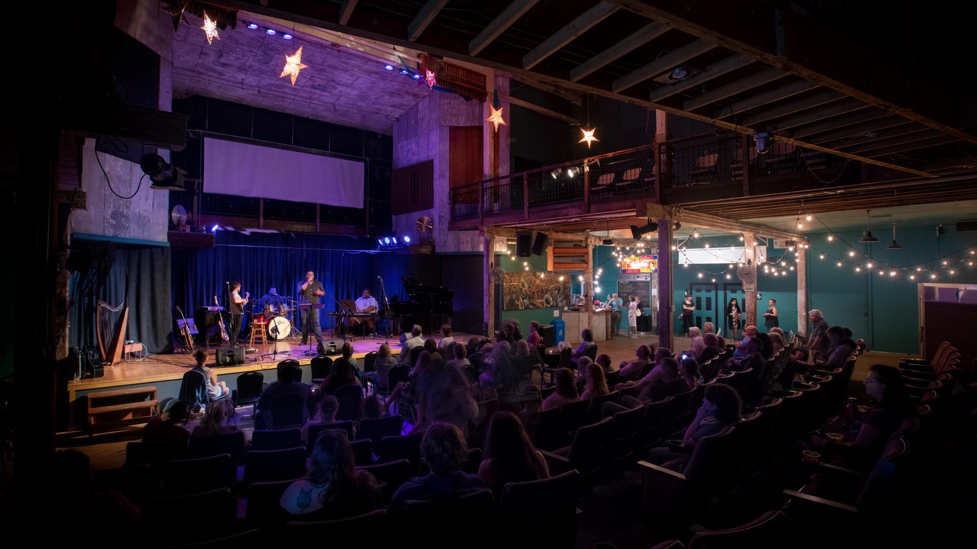 Inside Acorn Theater, an audience seated in rows watches a band perform under bright stage lights, while the space is softly lit by hanging stars and string lights.