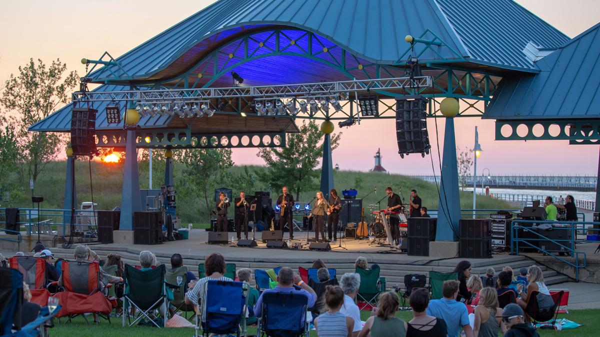 An audience sitting on lawn chairs and blankets outdoors in front of Shadowland Pavillion watches a jazz band perform on stage during a beach sunset.