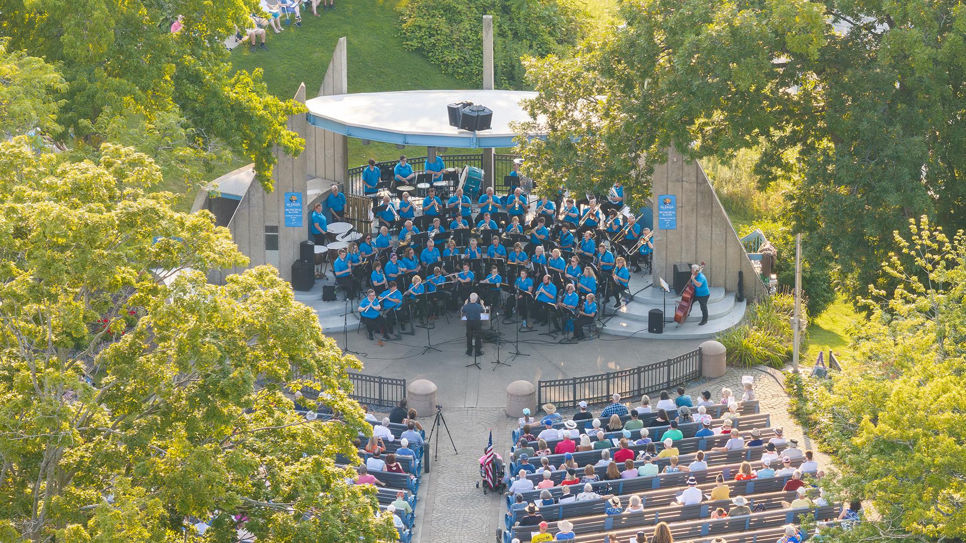 An aerial view of a large concert band performing outdoors on stage at John EN Howard Bandshell on a sunny day, an audience seated in rows watches.