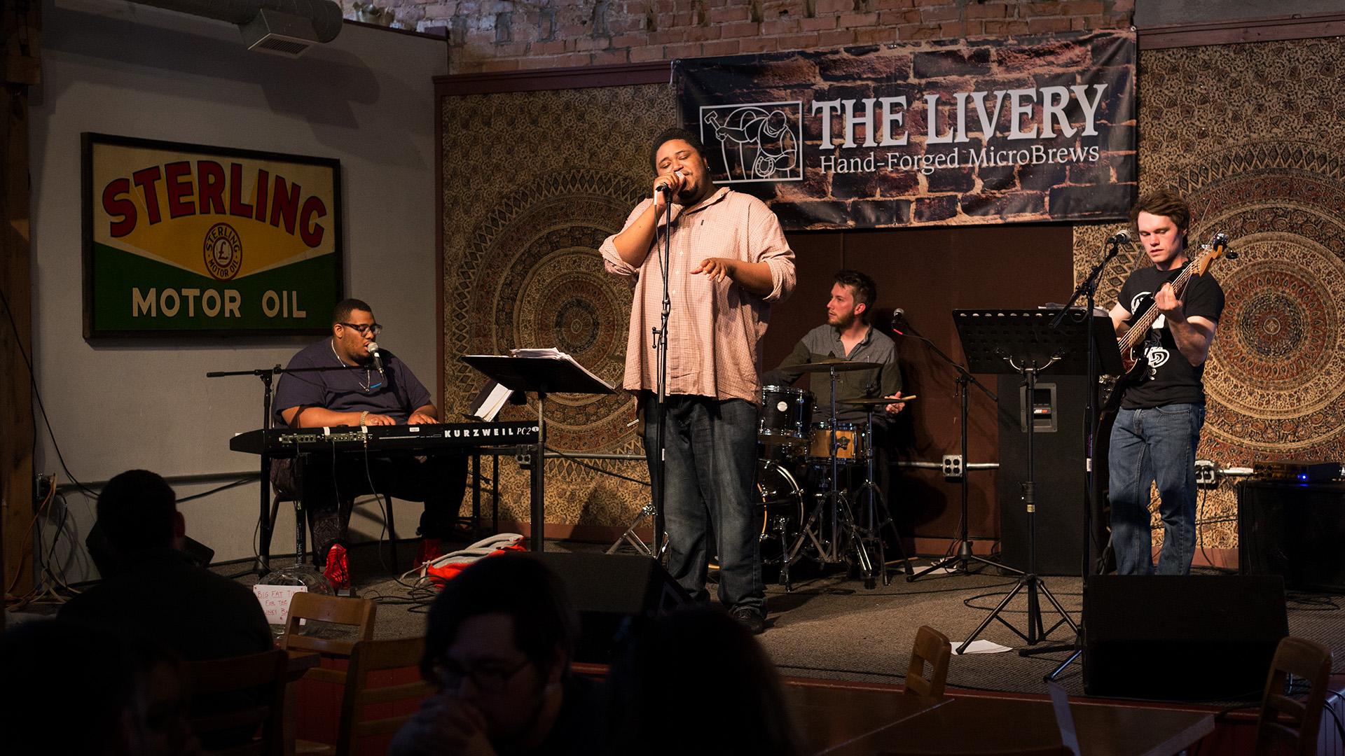 A band performs on stage at The Livery, a vocalist sings while band members play keyboard, drums, and bass guitar.
