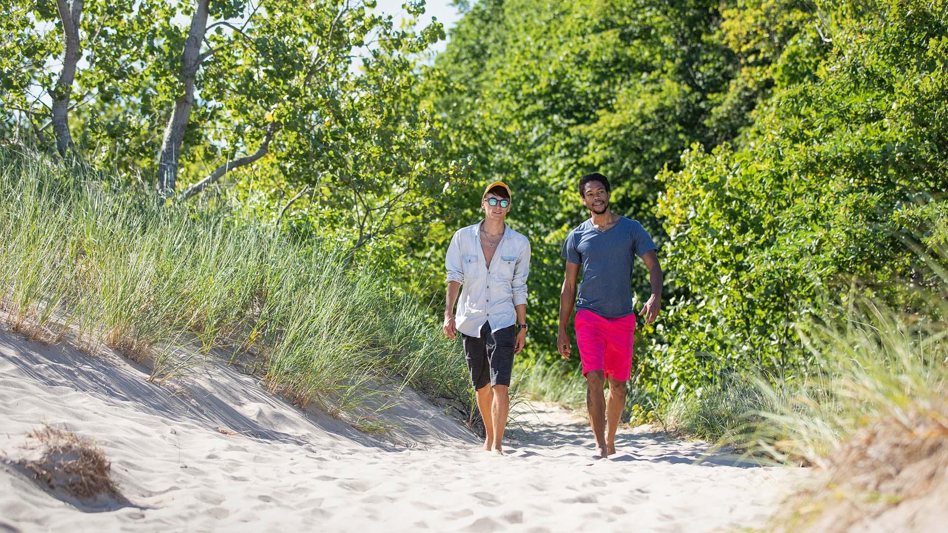 Two men walk barefoot on a sandy trail bordered by tall grass and trees on a sunny day at Grand Mere State Park.