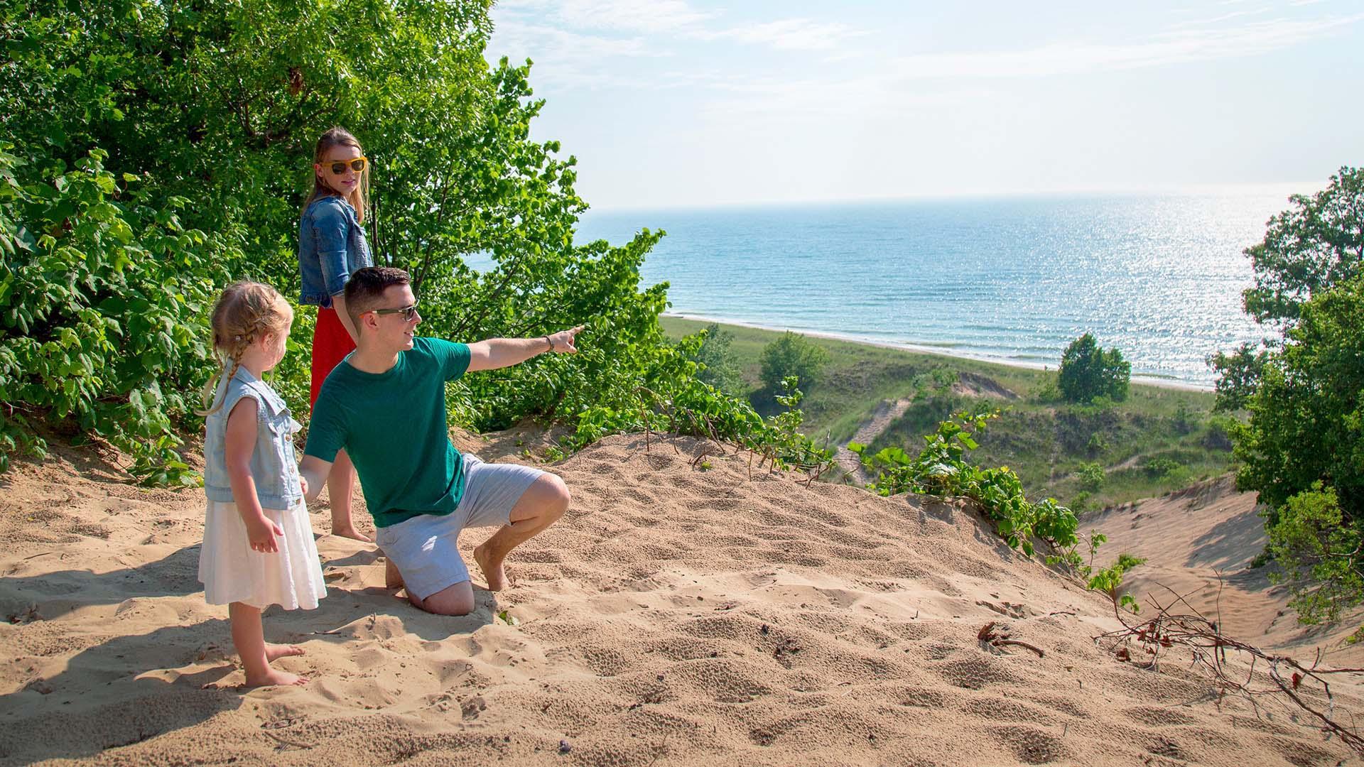 A man kneels in the sand atop a tall dune on a sunny day, gesturing toward the view of Lake Michigan for two children beside him at Grand Mere State Park.
