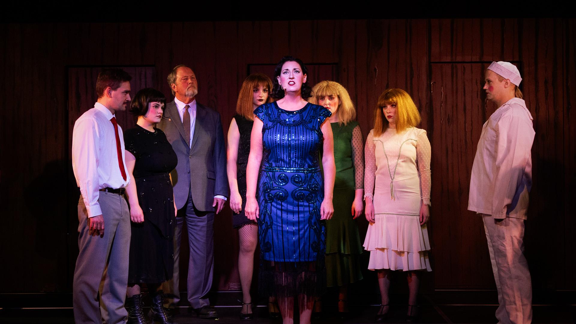A woman performer, brightly lit by a spotlight, is surrounded by costumed cast members on stage at GhostLight Theatre.