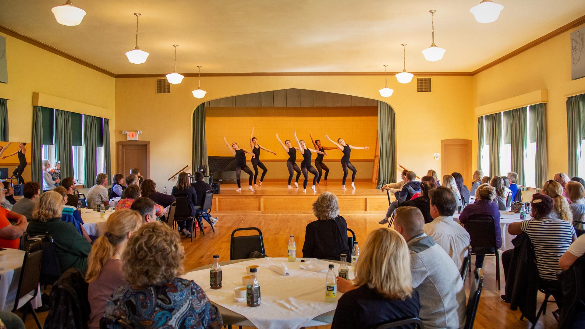 An audience seated at round tables watches a group of dancers perform in a brightly lit room at Citadel Dance & Music Center.