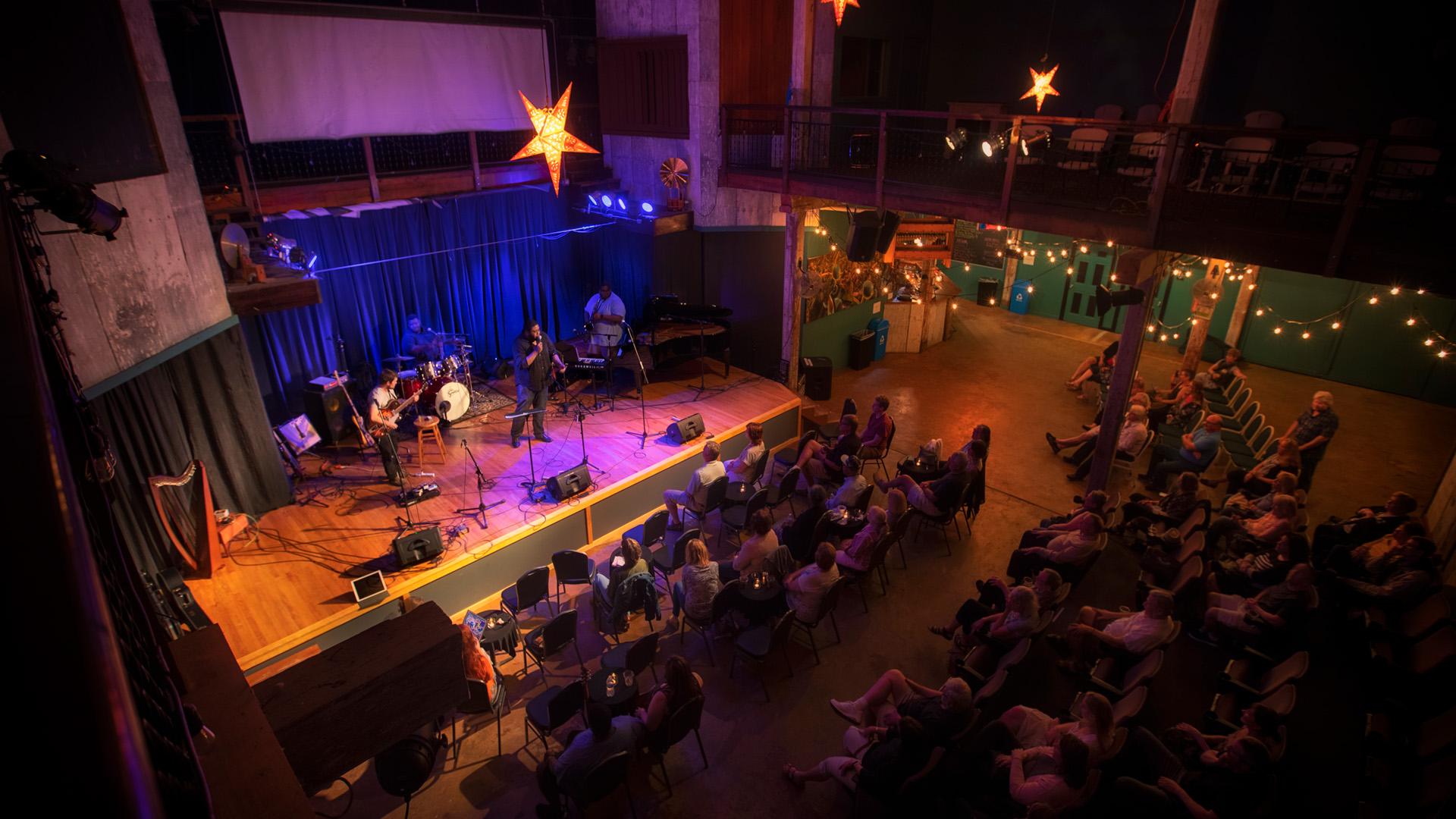 A top down view inside Acorn Theater, an audience seated in rows watches a band perform under stage lights, the space softly lit by hanging stars and string lights.