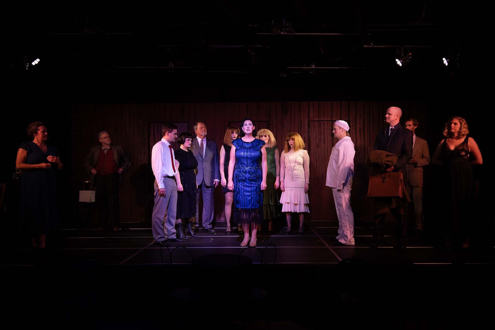 A woman performer, brightly lit by a spotlight, is surrounded by costumed cast members on stage at GhostLight Theatre