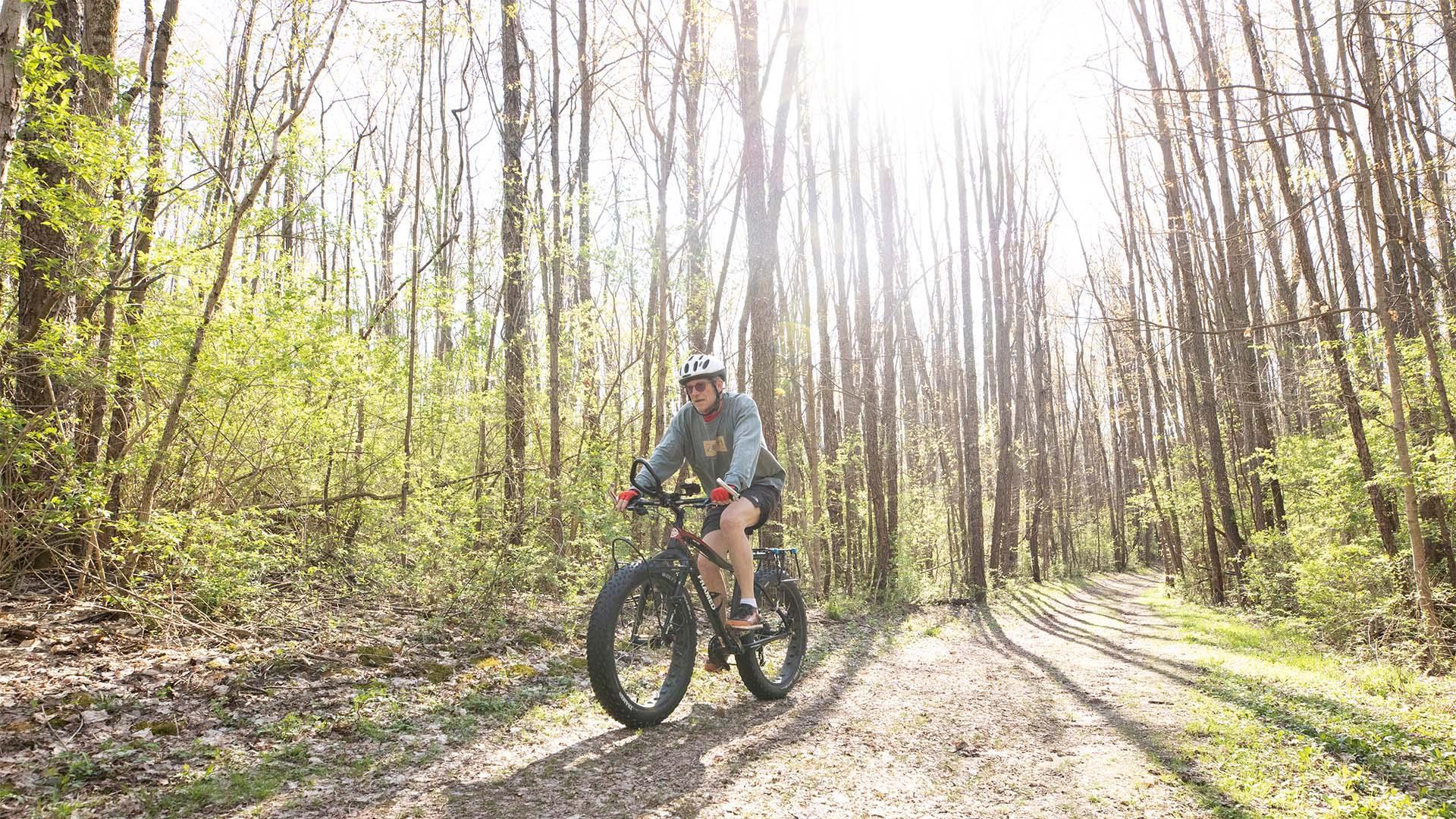 A person wearing a helmet rides a fat-tire bicycle on a sunlit gravel trail through a forest with tall, leafless trees at Love Creek County Park.