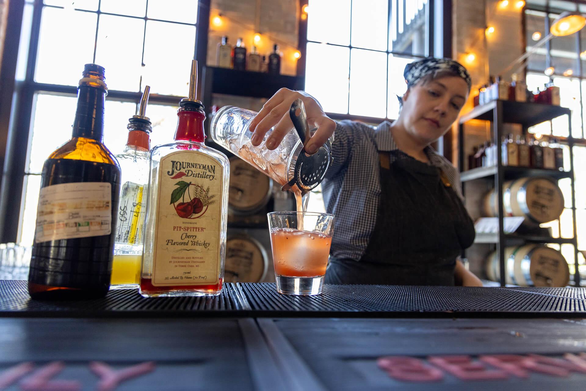 A bartender in a checkered shirt and apron pours a reddish-orange cocktail from a shaker into a glass at Journeyman Distillery. Whiskey bottles and barrels are in the background.