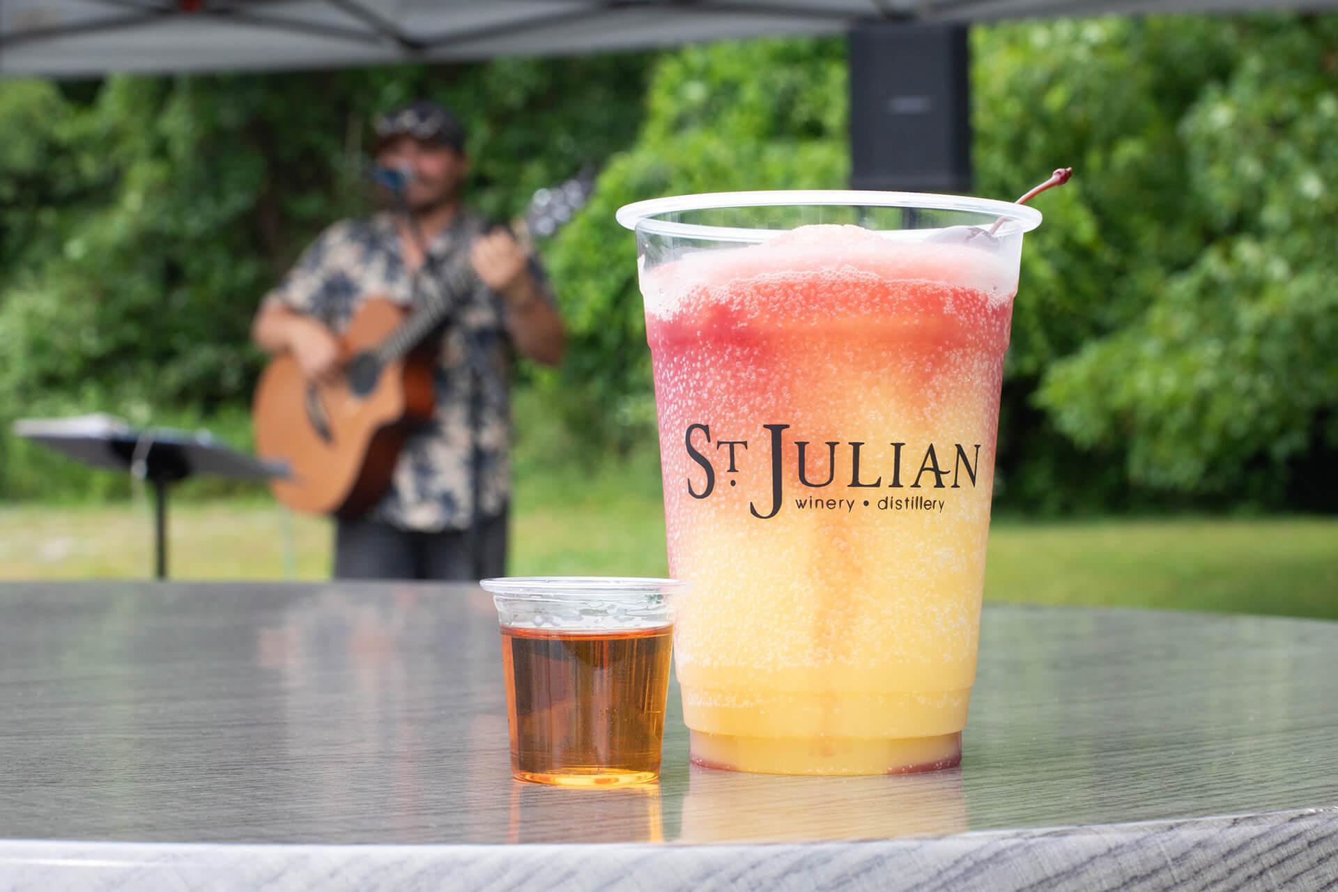 A colorful frozen drink and a small whiskey shot sit on a table, branded with -St. Julian Winery.- A musician plays guitar in the blurred background outdoors.
