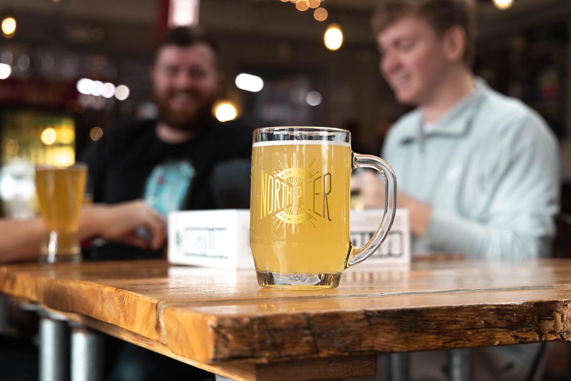 A glass of light beer with -NORTH PIER- written on it sits on a wooden table. Two people are in the background, smiling and chatting warmly in a cozy setting.