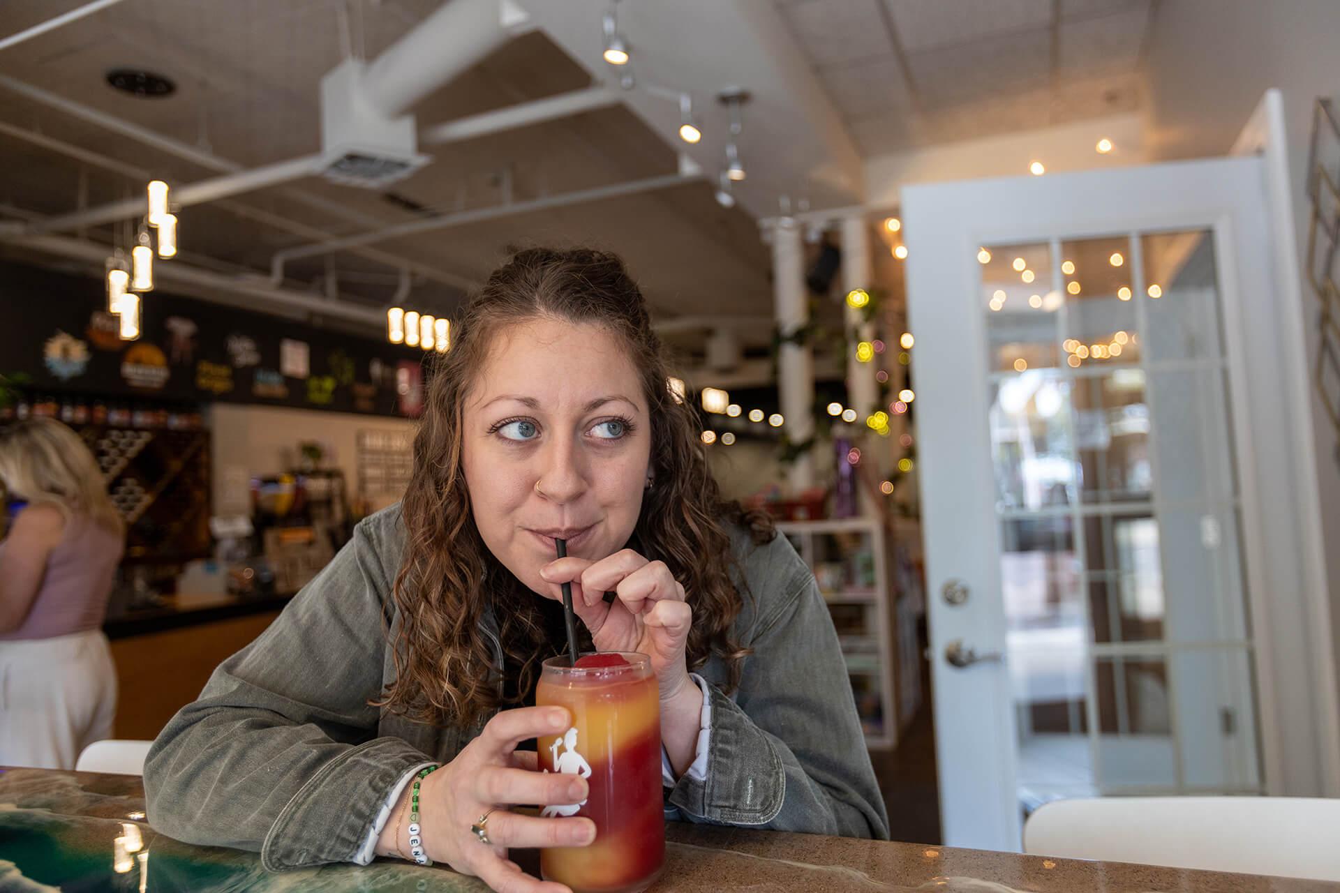 A woman with curly brown hair sips a colorful slushie through a straw at Lazy Ballerina Winery. She leans on the counter, looking content and relaxed.