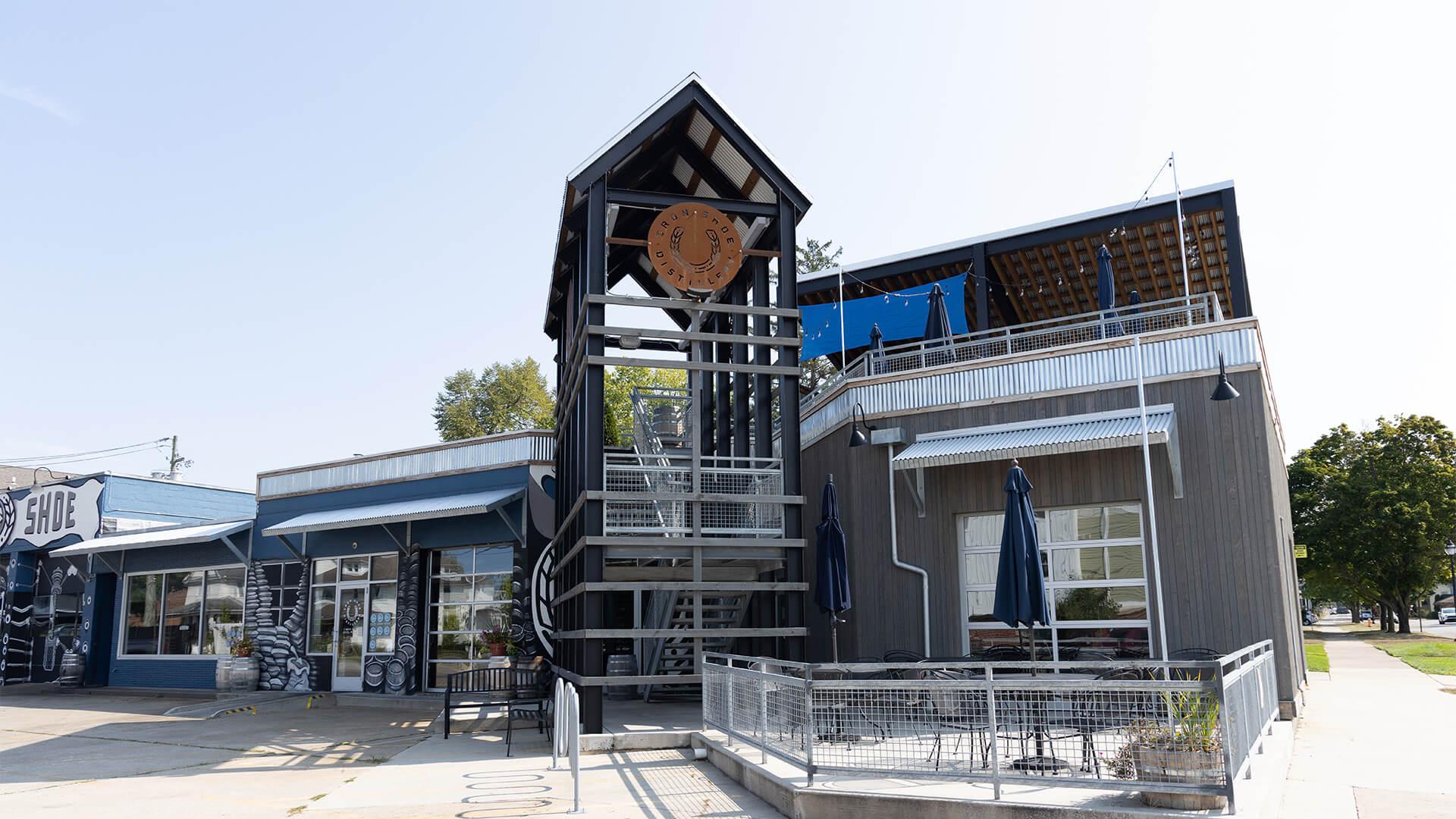 Iron Shoe Distillery is a modern building with a distinctive tower and spiral staircase stands on a sunny day. It features blue accents, large windows, and outdoor seating.