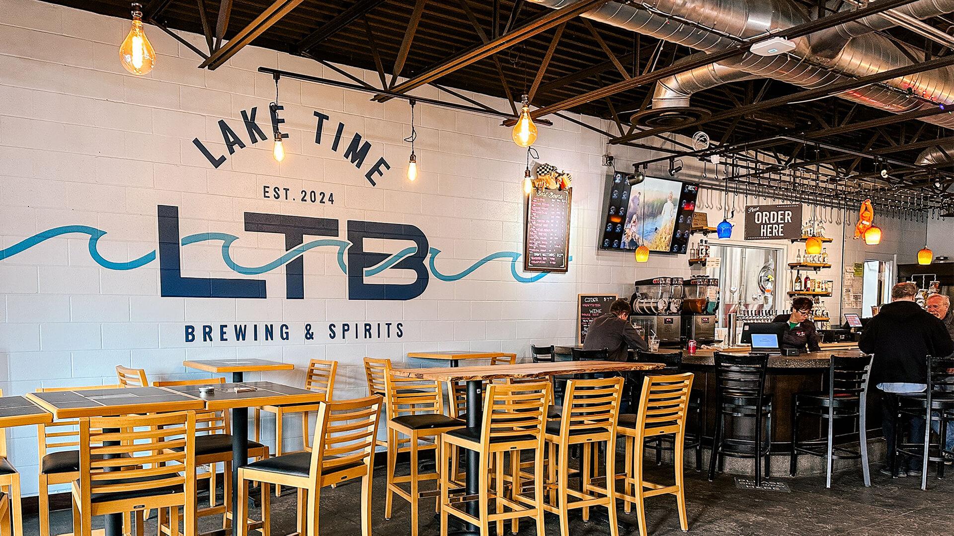 Lake Time Brewing & Spirits interior with wooden tables and chairs, industrial lighting, and a visible bar area where people are seated. Cozy, inviting atmosphere.
