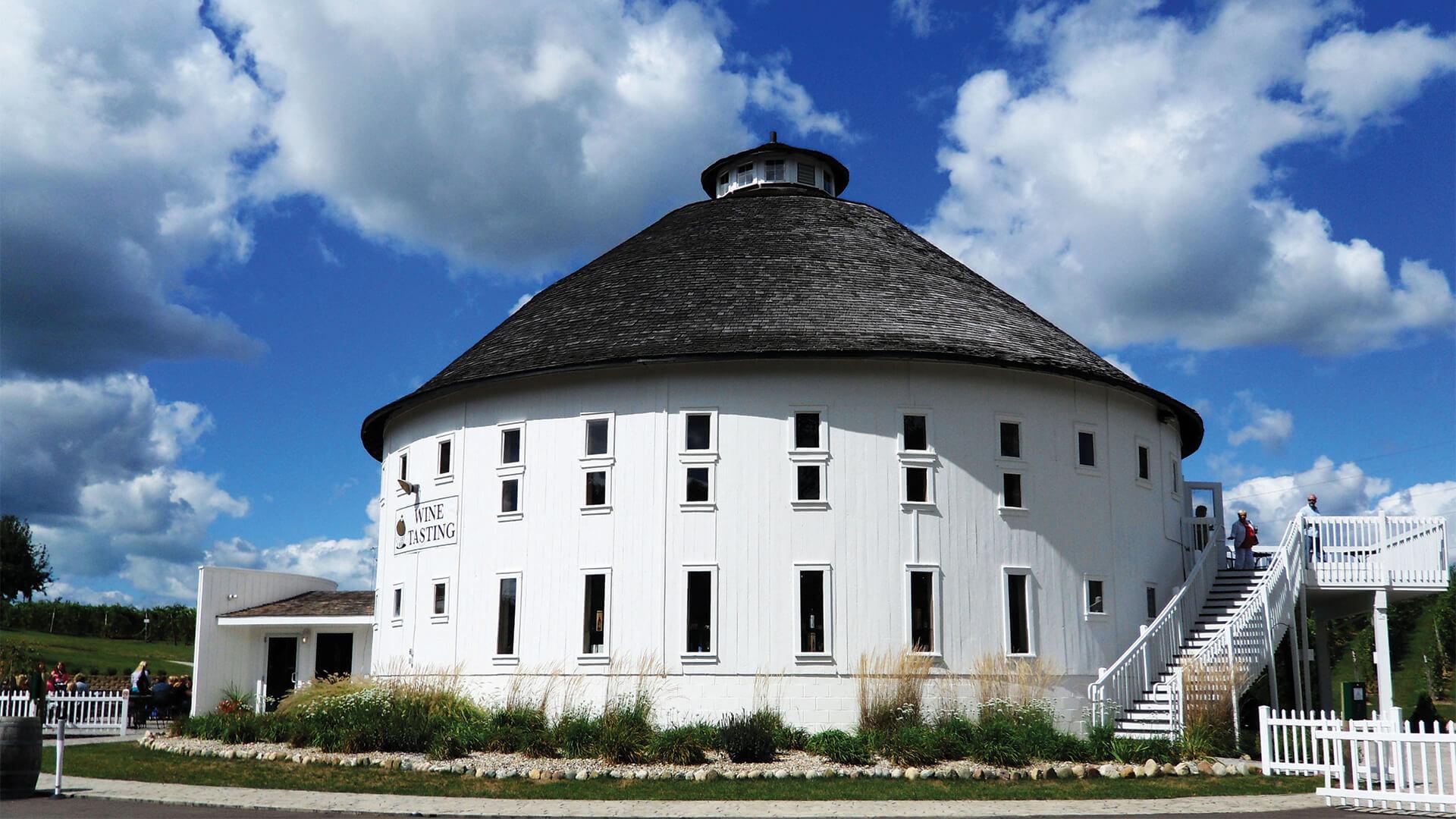 Round Barn Estate is a round white building with small windows and a dark conical roof against a blue sky with clouds. Sign reads -Wine Tasting.- People are near stairs.