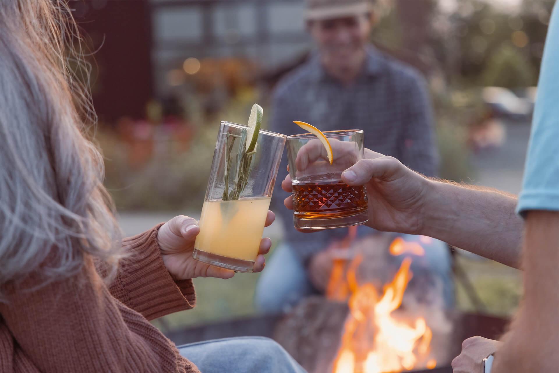 Two people clink glasses, one with a lemon cocktail and the other with a whiskey drink. A campfire glows in front, and a person sits blurred in the background.