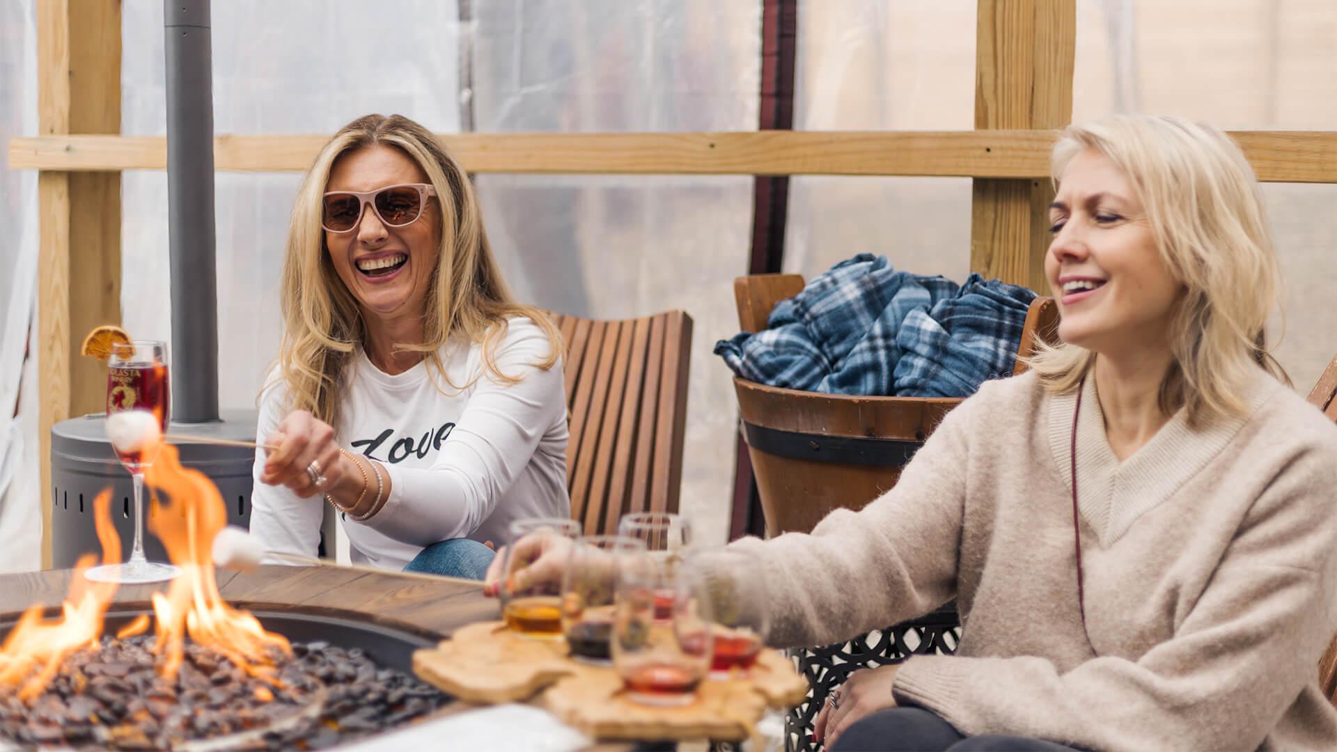Two women smile and roast marshmallows over a fire pit at Solasta Winery. Drinks and a cozy atmosphere suggest a relaxed and joyful outdoor gathering.