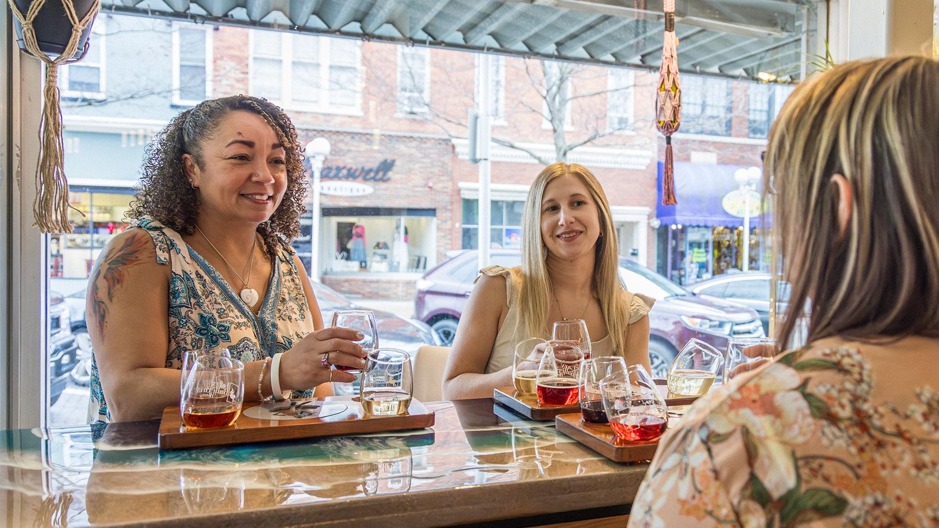 Three women sit at a bar, smiling and chatting, with tasting flights of wine in front of them. The atmosphere is warm and sociable, with a street view outside.