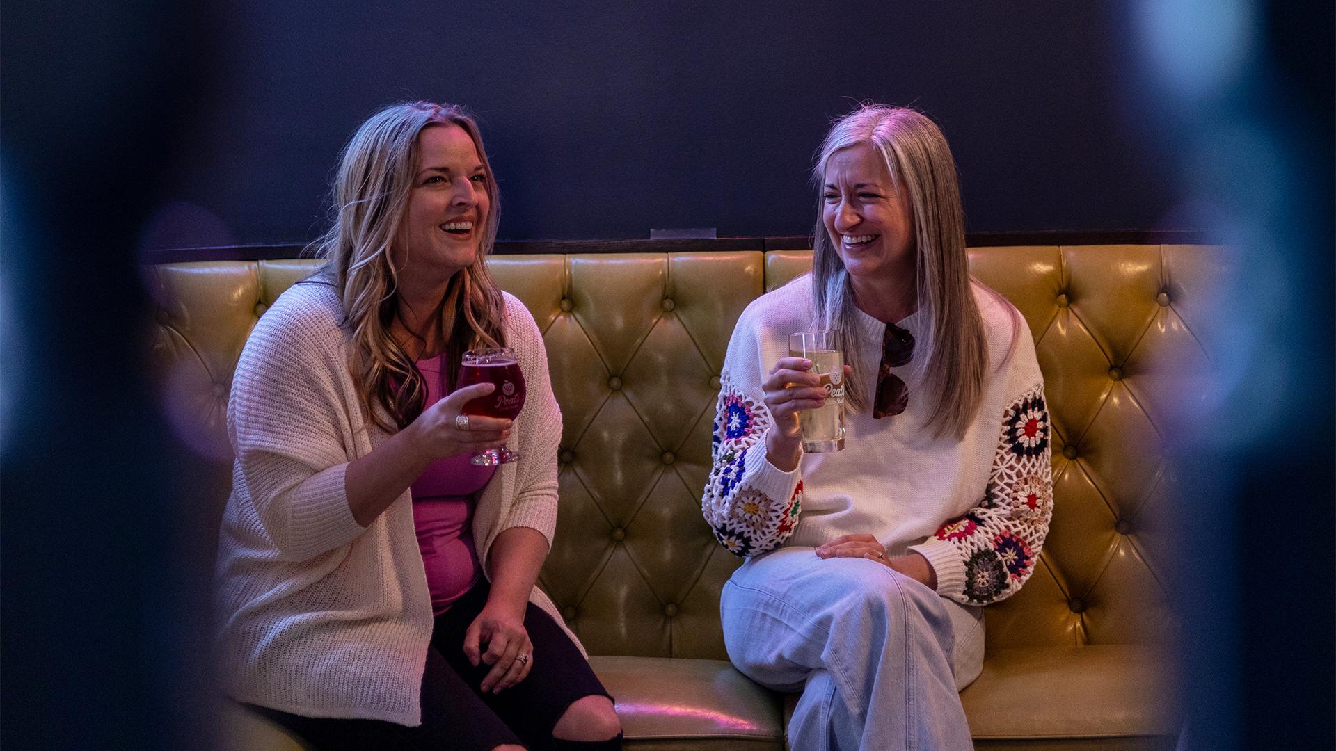 Two women sit on a yellow couch inside Peat