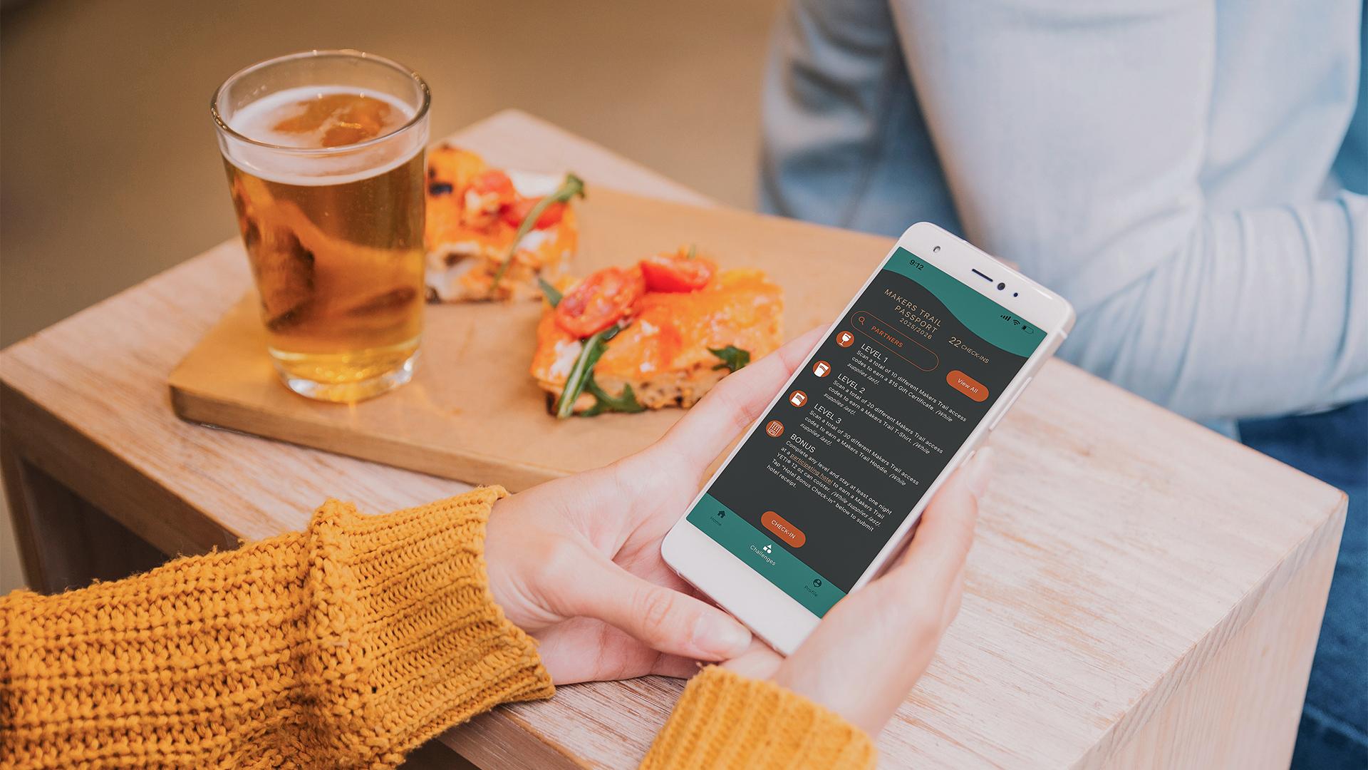 A person in a mustard sweater holds a smartphone displaying the Makers Trail app. Nearby, a wooden tray with tomato-topped bruschetta and a glass of beer sits on a table. The setting is cozy and casual.