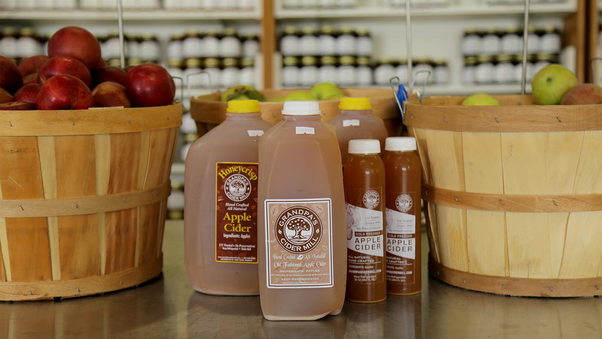Bottles of apple cider are displayed on a counter between two wooden baskets filled with red and green apples, set in a rustic market setting.