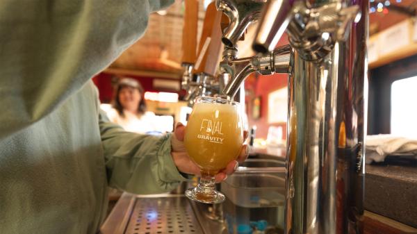 A person in a green shirt pours a craft beer labeled -Final Gravity- from a tap. The setting is cozy with warm lighting and a smiling bartender in the background.