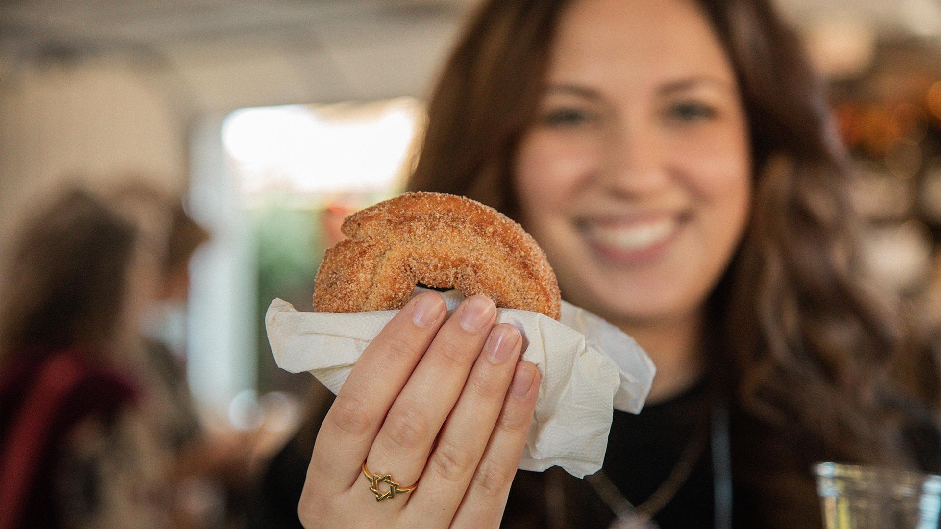 A smiling woman in a cozy setting holds up a cinnamon sugar pastry with a napkin. The mood is warm and inviting, emphasizing enjoyment and taste.