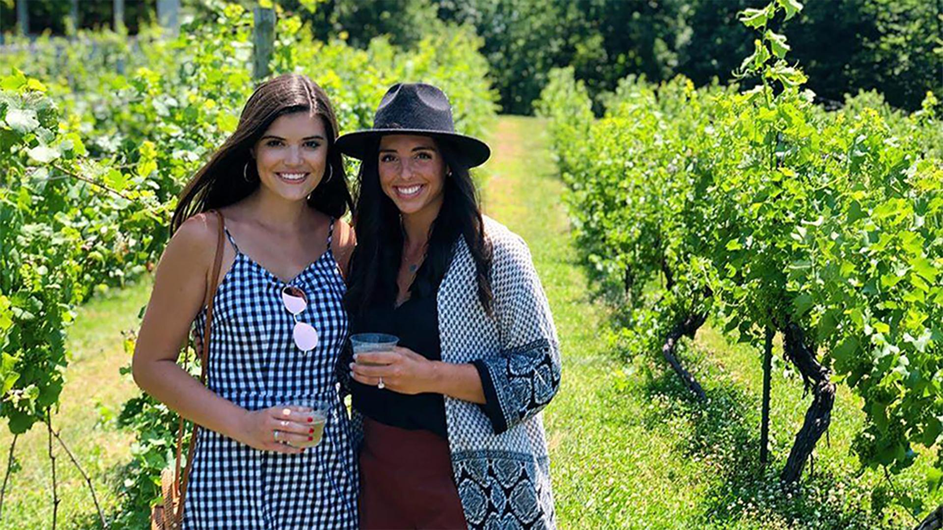 Two smiling women stand in a lush vineyard at Tabor Hill Winery under bright sunlight. One wears a checkered dress, the other a patterned cardigan and hat, both holding drinks.