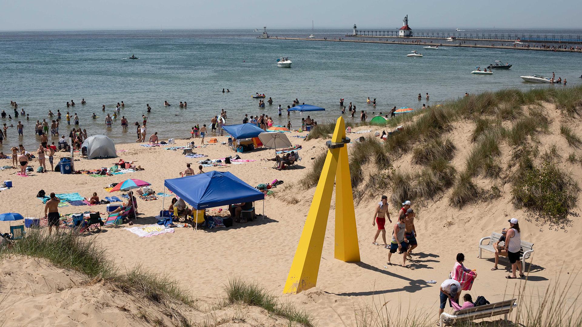 A busy afternoon at Silver Beach when the weather is hot. A large yellow sculpture in the foreground. In the background Lake Michigan, North Pier, boats, and the lighthouse.