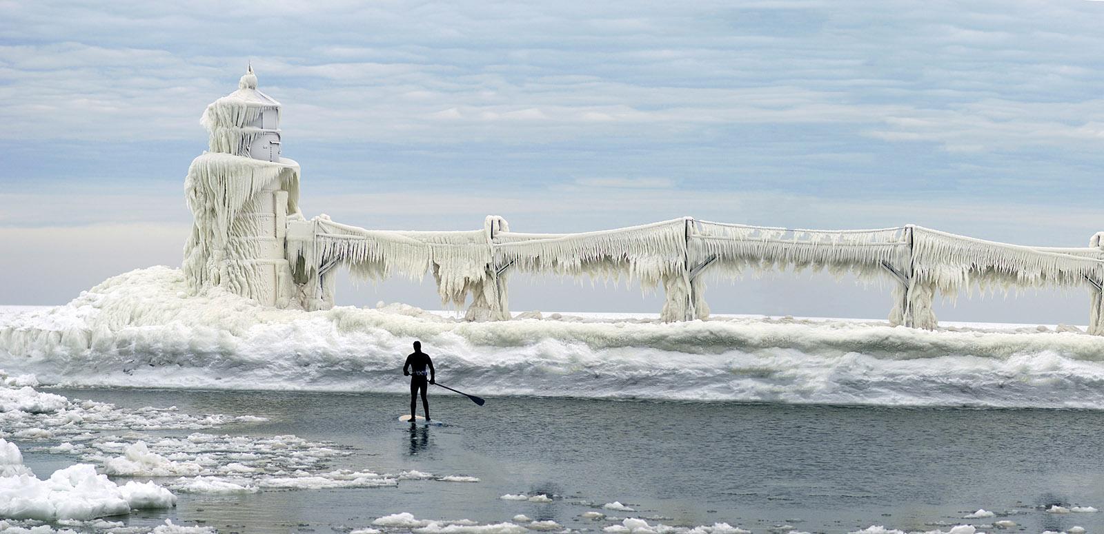A man stand-up paddleboarding in the St. Joseph River channel in winter, looking toward the ice-covered lighthouse.
