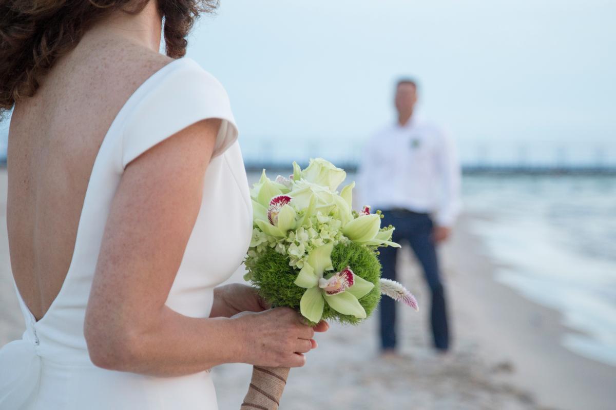 A bride walks along the beach holding flowers as she approaches the groom.